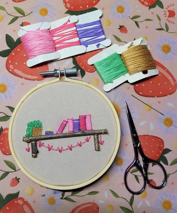 Embroidery hoop of a tiny bookshelf with pink books, beside floss and scissors